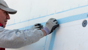 Ein Bauarbeiter mit Kappe und Handschuhen bringt ein blaues Armierungsgewebe auf weißen Dämmplatten an.