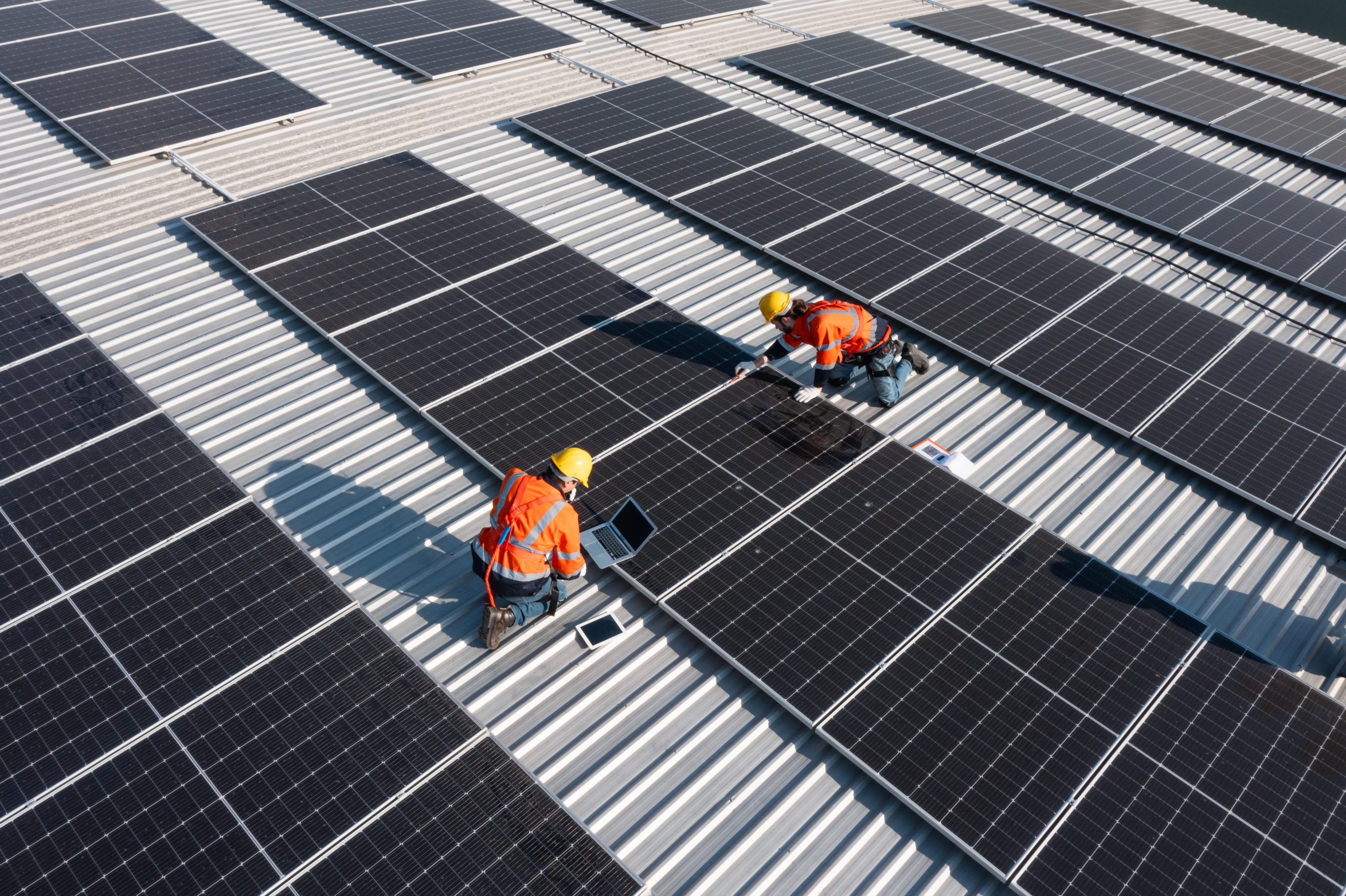 Zwei Techniker mit Schutzhelmen und Warnwesten bei Wartungsarbeiten auf einem mit Solarmodulen bedeckten Dach.